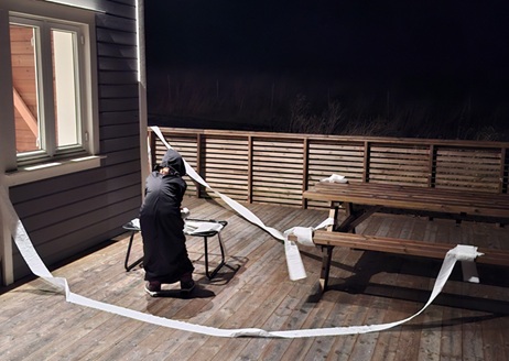 Vedkommende ankommer i langsomme, kontrollerte bevegelser. Personen stanser ved døren, tar fram en rull, og begynner å trekke papiret i stramme, diagonale linjer.